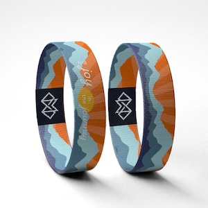 May include: Two elastic wristbands with a mountain and sun design in orange, blue, and teal. The bands have the text "Find joy in the journey" and a geometric logo on a black patch.