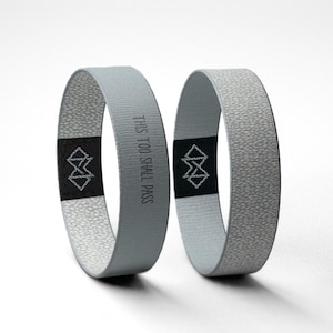 May include: Two light gray wristbands with a textured surface. One band has the text "THIS TOO SHALL PASS" printed on it. Both bands have a black patch with a white geometric design.