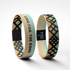 May include: Two woven wristbands with a geometric pattern in black, white, and teal. One wristband has the text "LOOK FOR THE GOOD" printed on it. The other wristband has the initials "MB" printed on it.