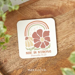 May include: A white coaster with a pink, orange, and green rainbow, two flowers, and the text "She is strong Proverbs 31:25".