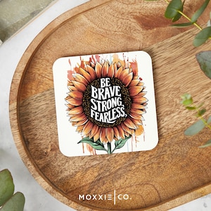 May include: A white coaster with a watercolor-style sunflower illustration. The sunflower has a black center with the text "BE BRAVE, STRONG, FEARLESS." The coaster has a brown wooden background.