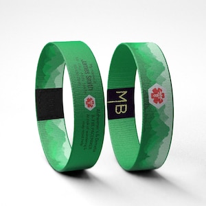 May include: Two green medical alert bracelets. One bracelet has the name "James Smith", a medical symbol, and contact information. The other bracelet has the initials "MB" and a medical symbol. Both bracelets have a mountain design.