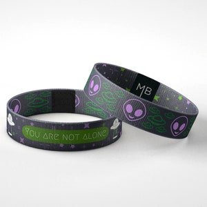 May include: Two black and green woven wristbands with a repeating pattern of alien faces and flying saucers. One wristband has the text "You are not alone" printed on it.