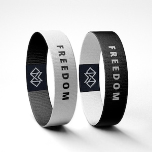 Puede incluir: Dos pulseras de tela, una blanca y otra negra, cada una con la palabra "FREEDOM" impresa verticalmente en blanco. Un logotipo geom&eacute;trico est&aacute; centrado en una etiqueta azul marino en cada pulsera. Las pulseras se muestran sobre un fondo blanco.
