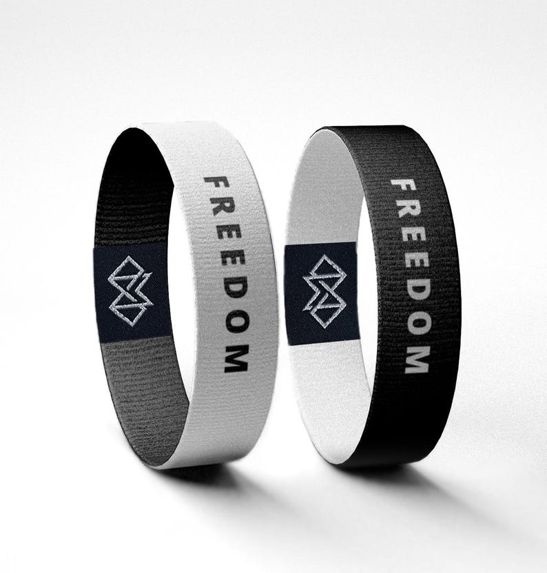 Puede incluir: Dos pulseras de tela, una blanca y otra negra, cada una con la palabra "FREEDOM" impresa verticalmente en blanco. Un logotipo geom&eacute;trico est&aacute; centrado en una etiqueta azul marino en cada pulsera. Las pulseras se muestran sobre un fondo blanco.