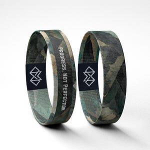 May include: Two camouflage pattern wristbands with the text "PROGRESS, NOT PERFECTION" in white. Each band has a black label with a white geometric design. The bands are made of a flexible material.
