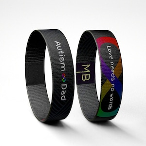 Könnte beinhalten: Zwei schwarze gewebte Armbänder mit weißem Text. Ein Band sagt "Autism Dad" mit einem Regenbogen-Unendlichkeitszeichen. Das andere Band sagt "Liebe braucht keine Worte" mit einem Regenbogen-Unendlichkeitszeichen.
