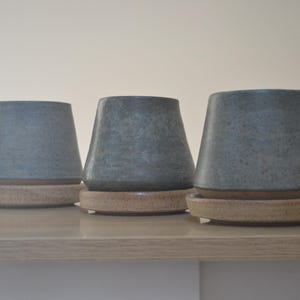 以下が含まれることがあります： 斑点のある青い釉薬と薄い茶色の縁のあるセラミックカップが3つ。各カップは、それに合う受け皿の上に置かれています。