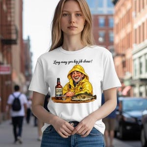 Puede incluir: Camiseta blanca con una ilustración de dibujos animados de un pescador con impermeable amarillo, una botella de licor, un vaso de chupito y un pez. El texto "Long may yer big jib draw!" está encima de la ilustración. La camiseta la lleva una persona.