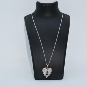 May include: A silver necklace featuring a pendant in the shape of angel wings. The wings are intricately detailed and seem to be set with small, sparkling stones. The necklace is displayed on a black mannequin.