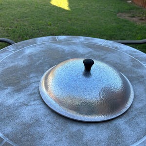 May include: A silver metal fire pit lid with a black handle sits on a gray stone table. The table is round and has a decorative tile pattern. The background shows green grass and a wooden fence.