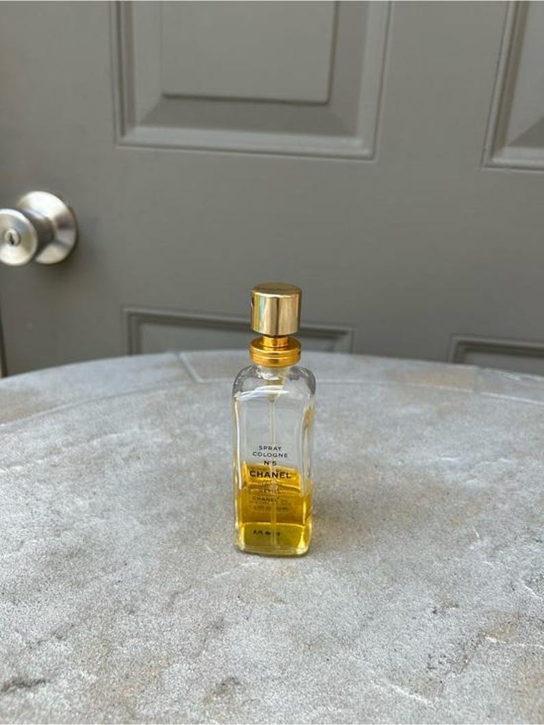 Puede incluir: Una botella de vidrio transparente de Chanel No. 5 Spray Cologne con tapa y detalles dorados. La botella es rectangular con bordes redondeados y contiene un l&iacute;quido de color dorado. La etiqueta es visible.