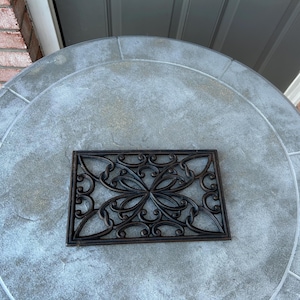 May include: A decorative, rectangular, dark brown metal mat with an ornate, symmetrical floral design. The mat is placed on a large, round, light gray stone table with a textured surface. The table is outdoors.