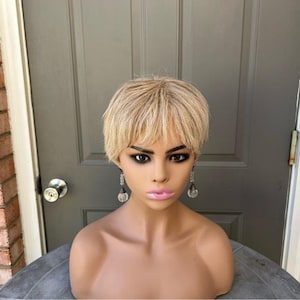 May include: A mannequin head with a short, blonde wig and dark eye makeup. The mannequin is wearing clear, dangling earrings. The background is a gray door and a brick wall.