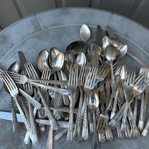May include: A collection of vintage silverware, including forks, spoons, and knives, arranged on a gray surface. The silverware is made of a shiny, silver-colored metal, with ornate designs on the handles of some pieces. The utensils are of various sizes.