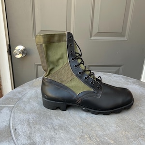 May include: A military-style combat boot featuring a black leather lower section and an olive green canvas upper shaft. The boot includes black laces, a series of metal eyelets for lacing, and a thick, lugged black rubber sole for traction.