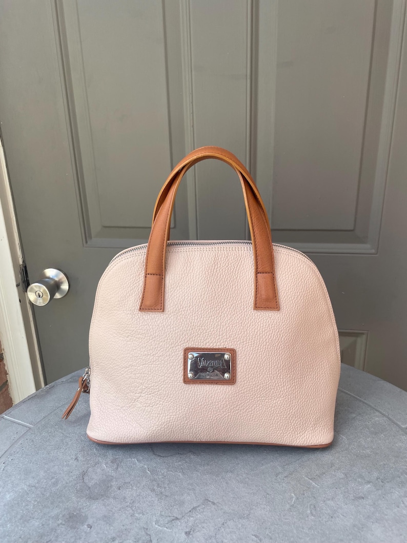 May include: A light pink leather handbag with brown leather handles and a silver metal nameplate that reads "Valentino."
