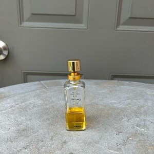 Puede incluir: Un peque&ntilde;o frasco de perfume rectangular con tap&oacute;n dorado y cuerpo de vidrio transparente. El frasco contiene un l&iacute;quido amarillo dorado. El nombre de la marca es visible en el frasco. El frasco est&aacute; sobre una superficie gris texturizada.
