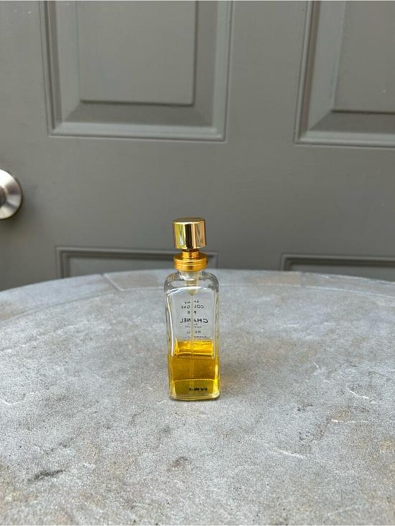Puede incluir: Un peque&ntilde;o frasco de perfume rectangular con tap&oacute;n dorado y cuerpo de vidrio transparente. El frasco contiene un l&iacute;quido amarillo dorado. El nombre de la marca es visible en el frasco. El frasco est&aacute; sobre una superficie gris texturizada.