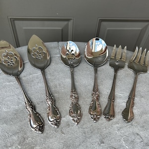 May include: A set of six silver-colored serving utensils, including spoons and forks, arranged on a gray surface. The spoons have decorative floral patterns on the bowl. The handles have an ornate, detailed design.