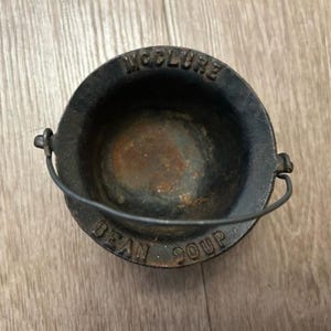 May include: A small, black cast iron pot with a handle. The pot has the words "McClure" and "Bean Soup" embossed on the rim. The pot is sitting on a light brown wooden surface.