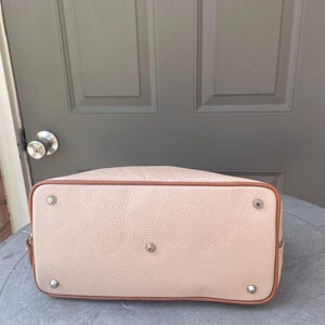 May include: A light pink leather handbag with a brown trim and silver hardware. The bag has a smooth texture and a slightly rounded shape.