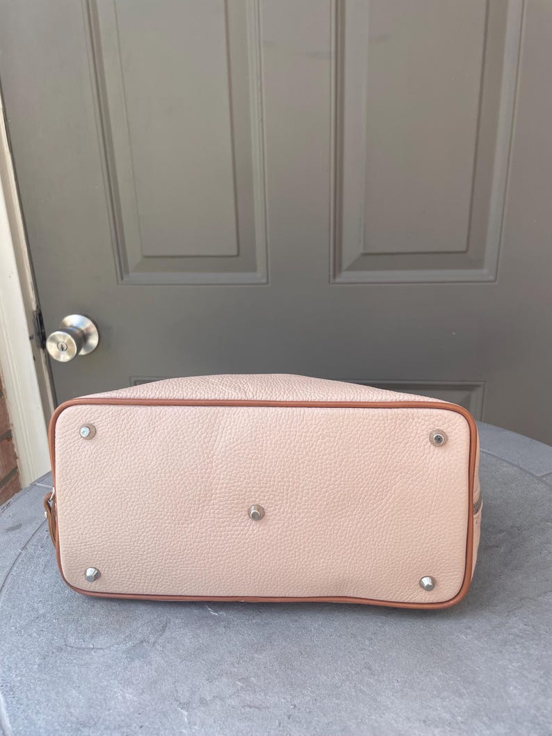 May include: A light pink leather handbag with a brown trim and silver hardware. The bag has a smooth texture and a slightly rounded shape.