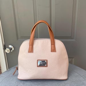 May include: A light pink leather handbag with a brown leather handle and a silver metal plate with the text "Valentino by Mario Valentino" and "Italy" on it.