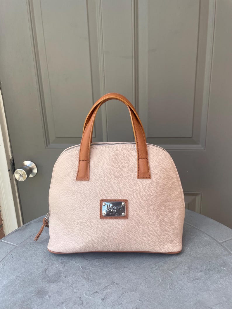 May include: A light pink leather handbag with a brown leather handle and a silver metal plate with the text "Valentino by Mario Valentino" and "Italy" on it.
