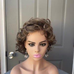 May include: A short, curly, brown wig with blonde highlights. The wig is styled with loose curls and is worn on a mannequin head. The mannequin is wearing silver earrings.