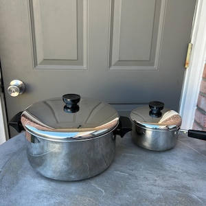 May include: Two stainless steel pots with black handles and lids. The larger pot is round with two side handles, and the smaller pot has a single handle. Both pots have black knobs on the lids. The pots are sitting on a gray surface.