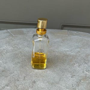 Puede incluir: Una botella de vidrio transparente de Chanel No. 5 Spray Cologne con tapa y detalles dorados. La botella contiene un l&iacute;quido amarillo y tiene una etiqueta con el nombre de la marca y los detalles del producto. La botella de colonia est&aacute; sobre una superficie gris.