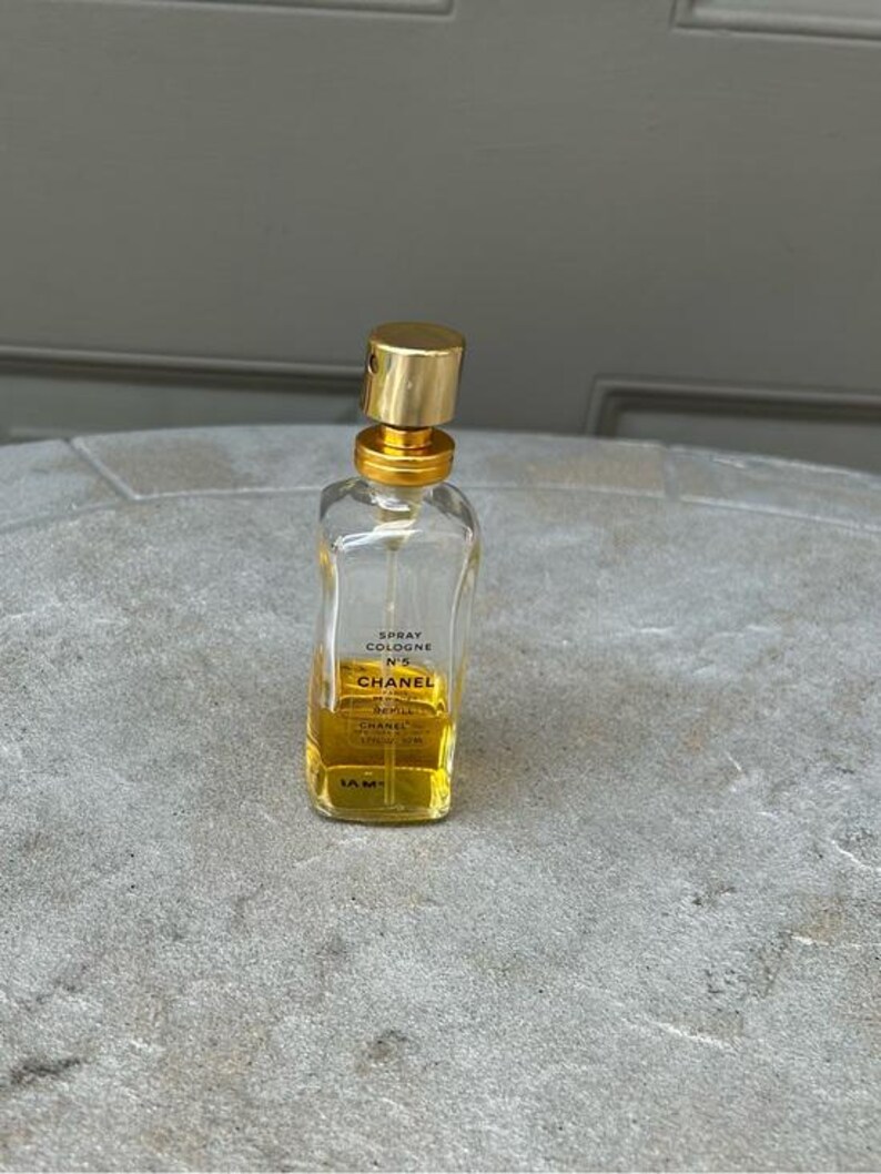 Puede incluir: Una botella de vidrio transparente de Chanel No. 5 Spray Cologne con tapa y detalles dorados. La botella contiene un l&iacute;quido amarillo y tiene una etiqueta con el nombre de la marca y los detalles del producto. La botella de colonia est&aacute; sobre una superficie gris.
