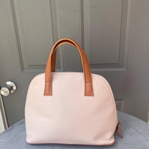 May include: A light pink leather handbag with brown leather handles. The bag has a dome shape and a zipper closure.