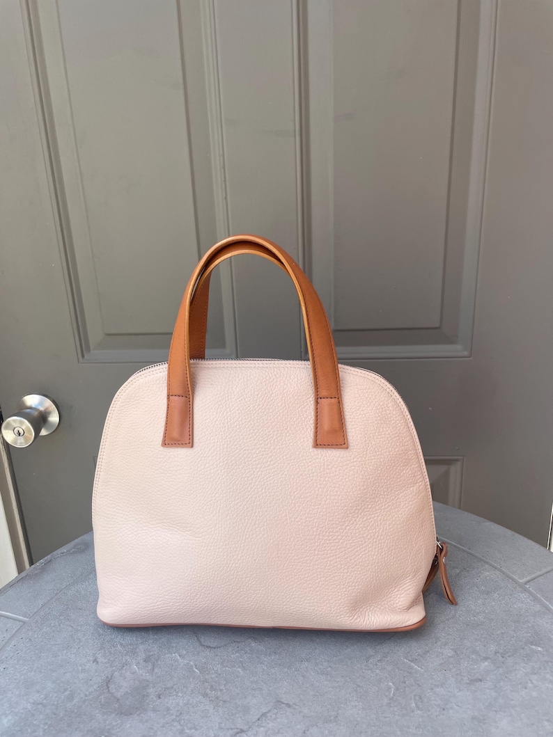 May include: A light pink leather handbag with brown leather handles. The bag has a dome shape and a zipper closure.