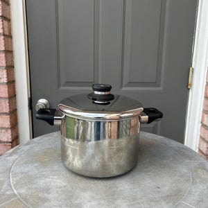 May include: A stainless steel stockpot with a polished finish, featuring two black side handles and a matching lid with a black knob. The pot has a cylindrical body and a flat base, designed for stovetop cooking with a clean, metallic appearance.