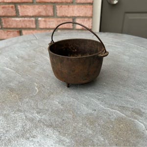 May include: A small, antique cast iron cauldron with a handle. The cauldron is dark brown with three small legs and a curved handle. The pot is sitting on a gray stone surface, with a brick wall in the background.