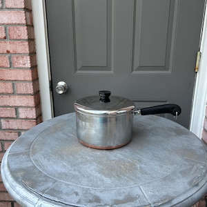 May include: A stainless steel saucepan with a black handle and lid, sitting on a round, gray stone table. The pot has a copper-colored bottom and is positioned in front of a gray door.