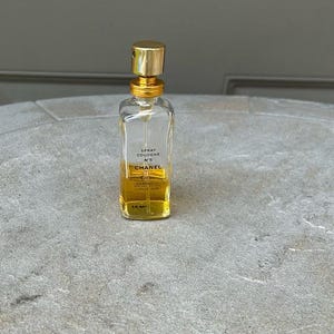 Puede incluir: Un frasco de perfume rectangular transparente con tap&oacute;n y detalles dorados. El frasco contiene un l&iacute;quido amarillo y tiene el texto "SPRAY COLOGNE N&deg;5 CHANEL" impreso en la parte delantera. El frasco est&aacute; sobre una superficie gris.