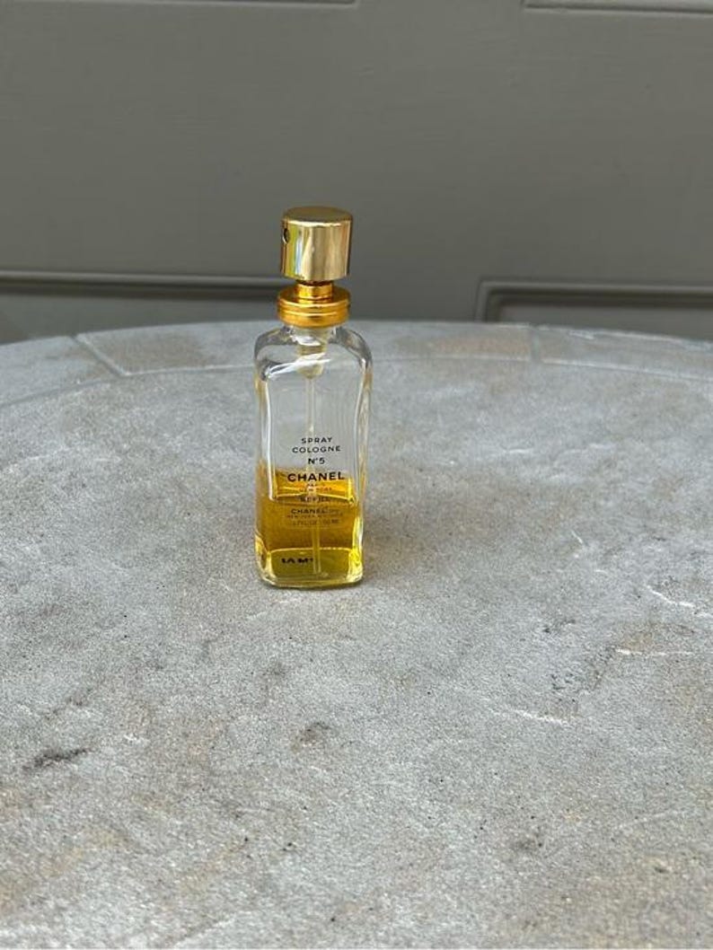 Puede incluir: Un frasco de perfume rectangular transparente con tap&oacute;n y detalles dorados. El frasco contiene un l&iacute;quido amarillo y tiene el texto "SPRAY COLOGNE N&deg;5 CHANEL" impreso en la parte delantera. El frasco est&aacute; sobre una superficie gris.