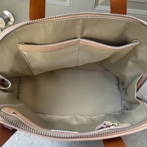 May include: A light pink leather tote bag with a zipper closure and two brown leather handles. The bag has a large interior compartment and two smaller pockets on the inside.