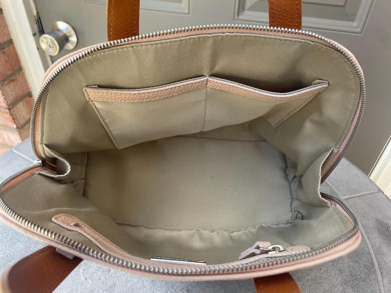 May include: A light pink leather tote bag with a zipper closure and two brown leather handles. The bag has a large interior compartment and two smaller pockets on the inside.