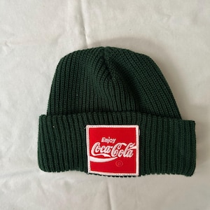 May include: A dark green knit beanie with a red Coca-Cola logo patch. The patch features the words "Enjoy Coca-Cola" in white lettering. The beanie is made of a ribbed knit material.