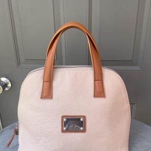 May include: A light pink leather handbag with brown leather handles and a silver metal nameplate that reads "Valentino Design".