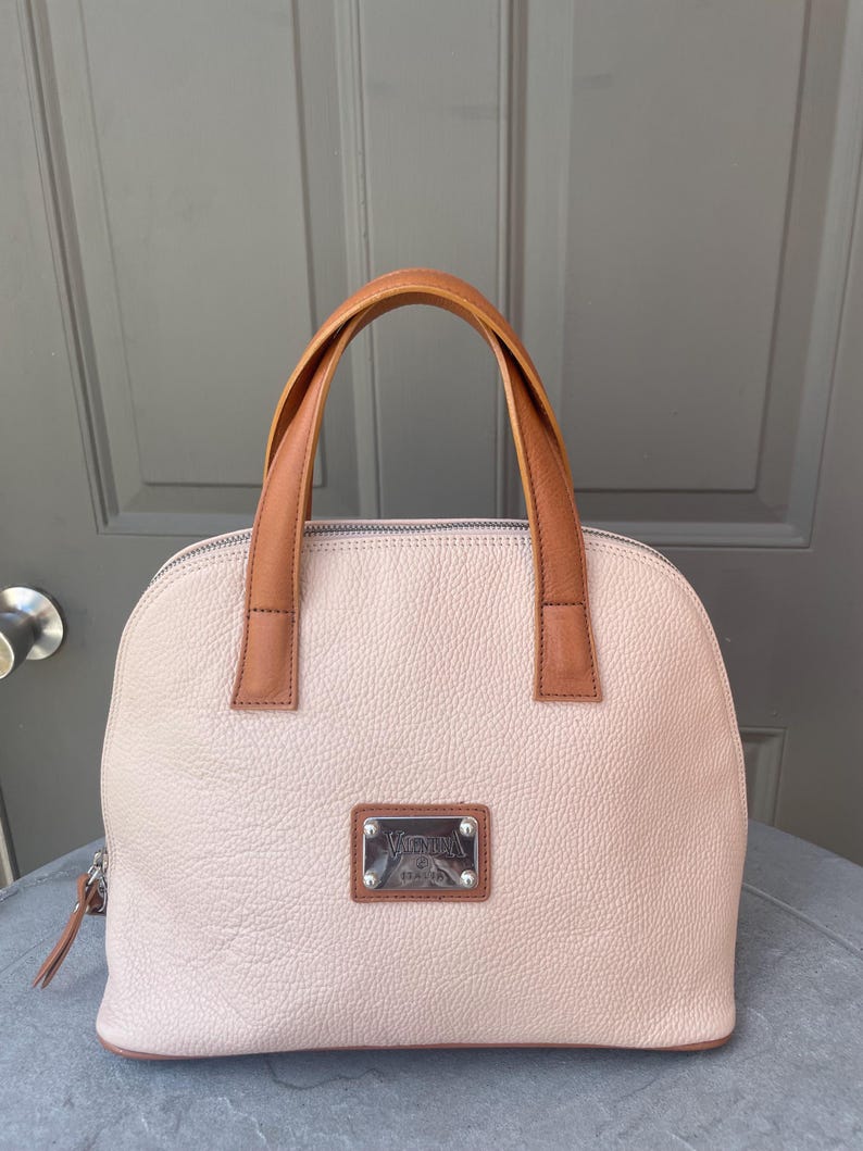 May include: A light pink leather handbag with brown leather handles and a silver metal nameplate that reads "Valentino Design".