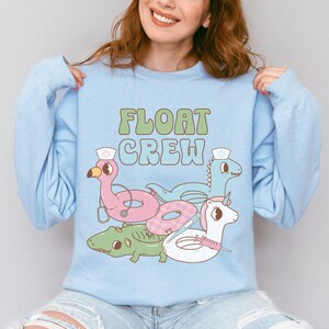 May include: Light blue sweatshirt with the text "FLOAT CREW" in green letters. The sweatshirt features a cartoon illustration of a pink flamingo, a blue dinosaur, a green alligator, and a unicorn, all wearing nurse hats and riding on inflatable pool floats.