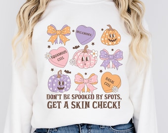 May include: White sweatshirt with a Halloween-themed design featuring pumpkins, bows, and hearts with the text "SQUAMOUS CELL", "MELANOMA", "BASAL CELL", and "DON'T BE SPOOKED BY SPOTS, GET A SKIN CHECK!"