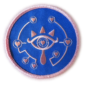 May include: A blue and pink embroidered patch featuring a stylised eye with hearts around it. The eye is surrounded by a circle of hearts and a line of hearts.