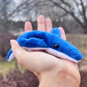 May include: A plush blue whale toy with white accents and a black eye. The whale is made of soft, fuzzy material and is held in a hand. The toy is a whimsical representation of a whale.
