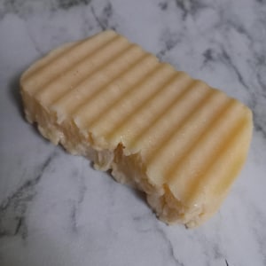 May include: A rectangular bar of light yellow soap with a ridged top. The soap has a slightly uneven texture on the sides and bottom. The soap is sitting on a marble surface.